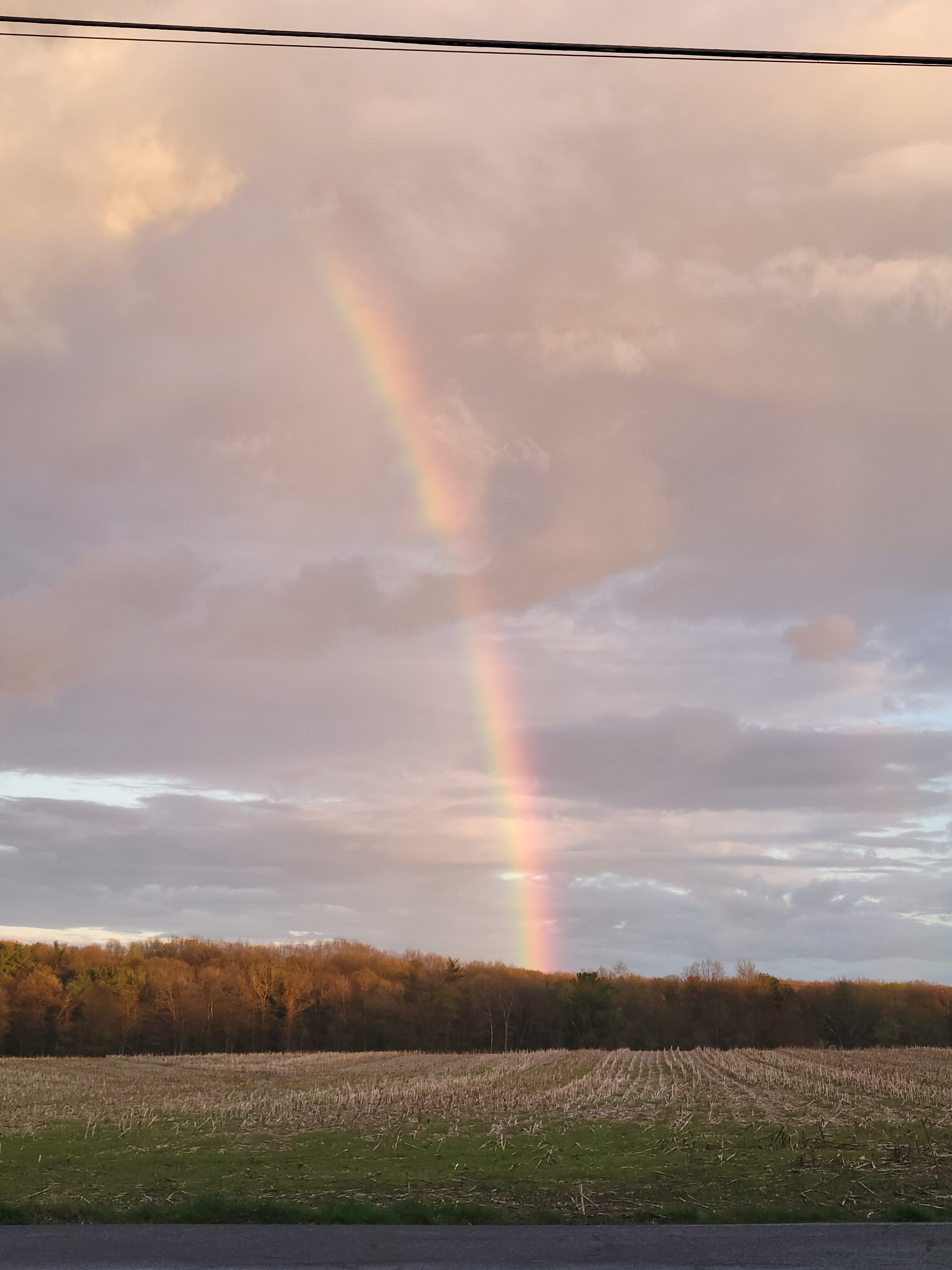 A rainbow shines through the dark clouds, casts itself over a barren field.