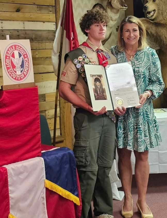 Sen. Rosemary Brown presents a Citation the newest Eagle Scout of Troop 84, Alexsander Tsyganov Lamberton. They are posing in front of the American Flag, as well as the Scout of Honor sign in the background.